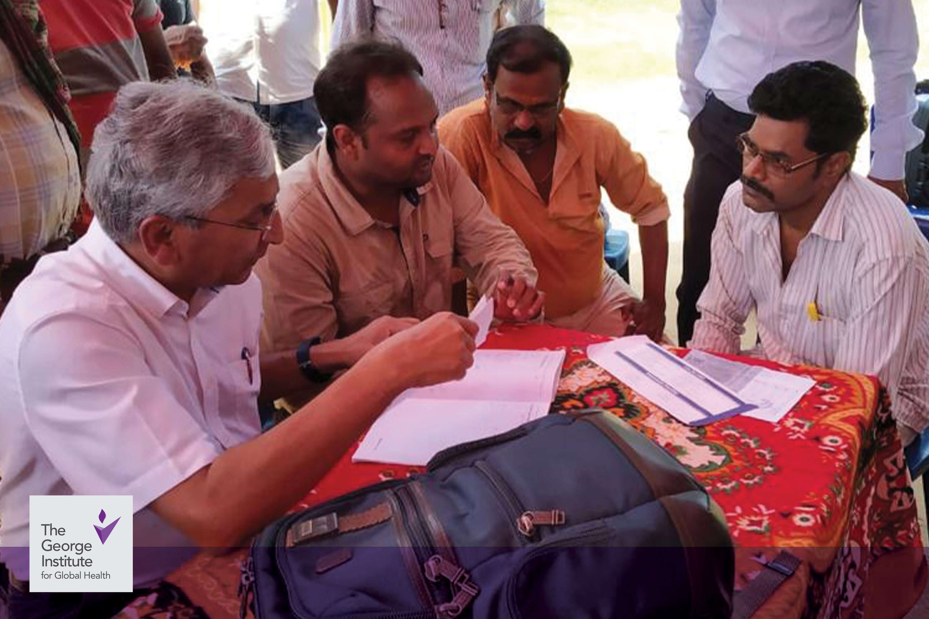 Four men sit at a table engaged in conversation; one is holding a piece of paper