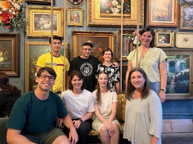 Eight people smile at the camera in two rows, in front of wall-to-wall paintings in ornate frames.