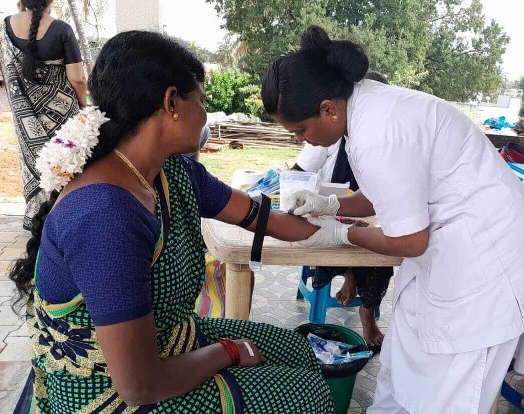 A female doctor takes a blood sample from a woman, sitting, dressed in a green, blue and yellow sari. A female assistant in a white coat sits on the other side of the table