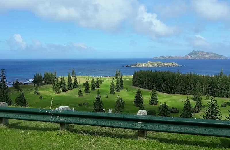 A landscape view of the Norfolk Island coast: bright green in the foreground with fir trees meet a bright blue ocean with two islands visible.
