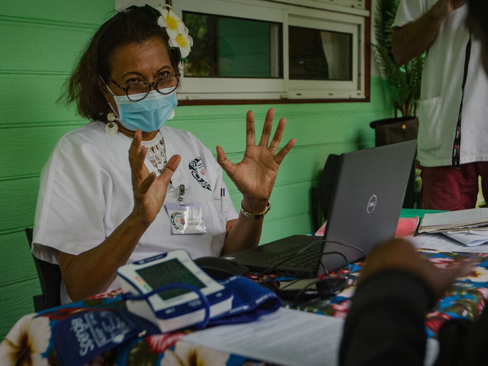 A woman sitting at a table wearing a face mask, with a blood pressure reader and laptop.