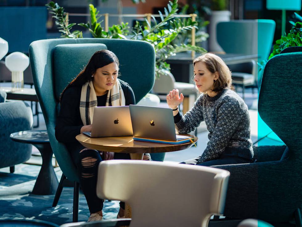 Two people sitting in an office with laptops on a table next to each other.