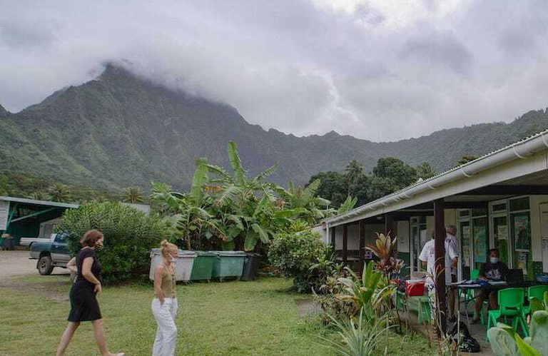Sample collection in Moorea, French Polynesia. Photo credit: Tom Martienssen Two women walk up to a single-story house surrounded by leafy vegetation. Clouds obscure a mountain rising behind them.