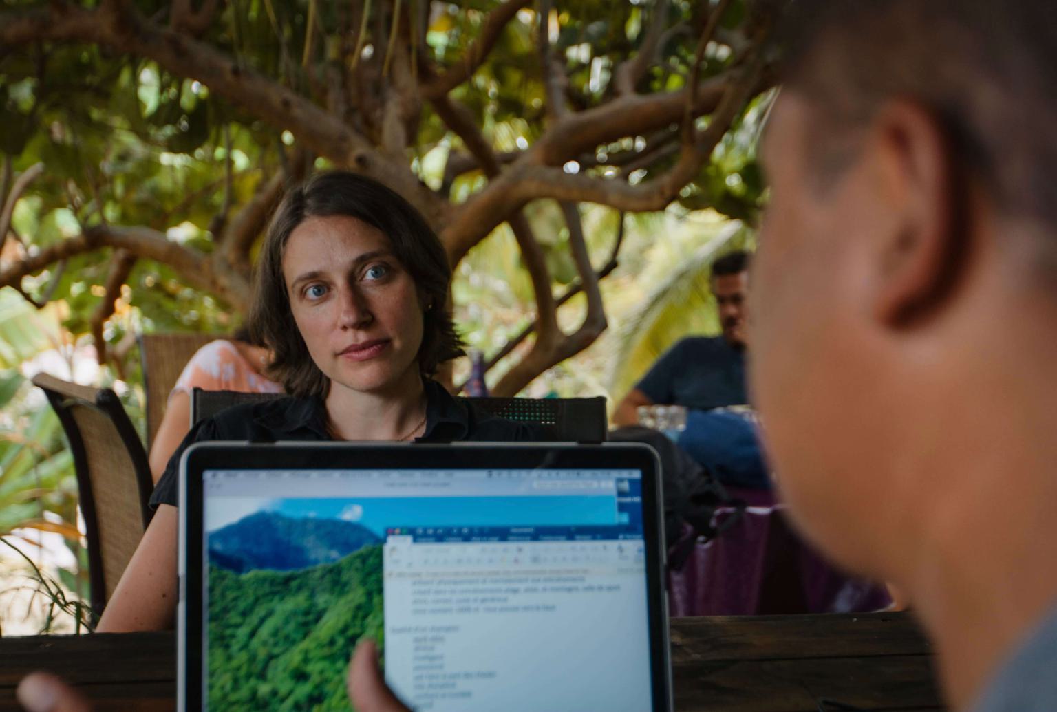 A man in the foreground in front of a computer. The photo looks over the computer at a woman across the table, listening to the man