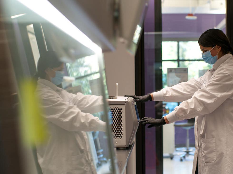 A scientist standing in a lab coat and face mask, operating lab equipment on a table.