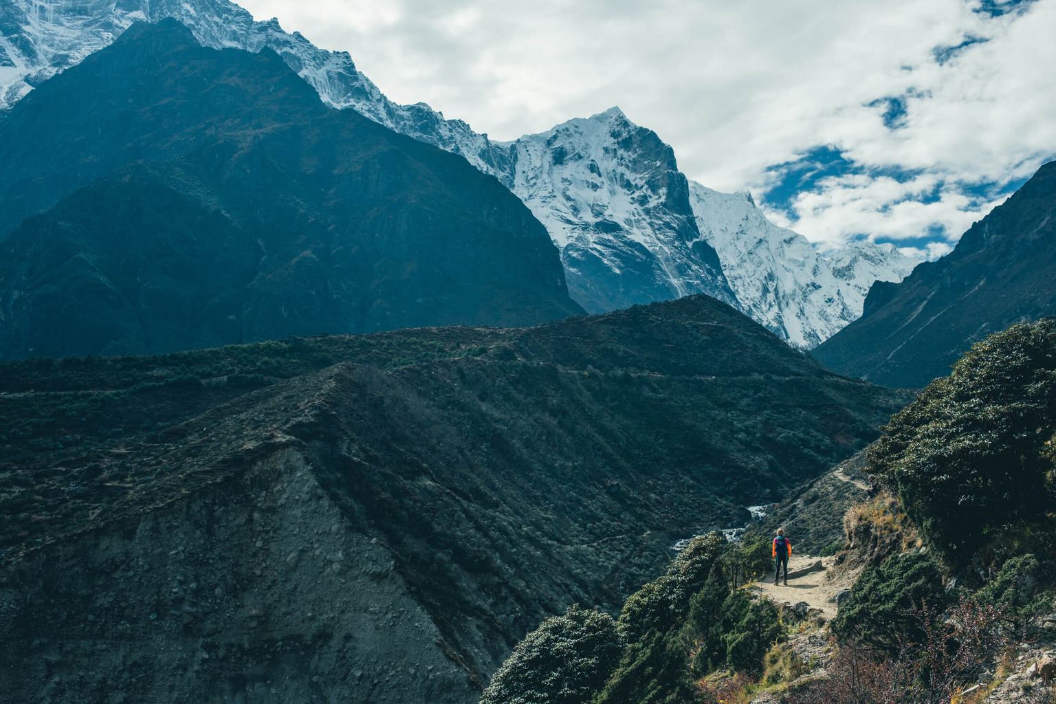 A hiker on a mountain trail with snow-capped mountains in the background.