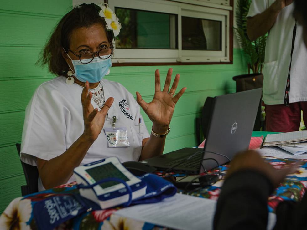 A woman sitting at a table wearing a face mask, with a blood pressure reader and laptop.